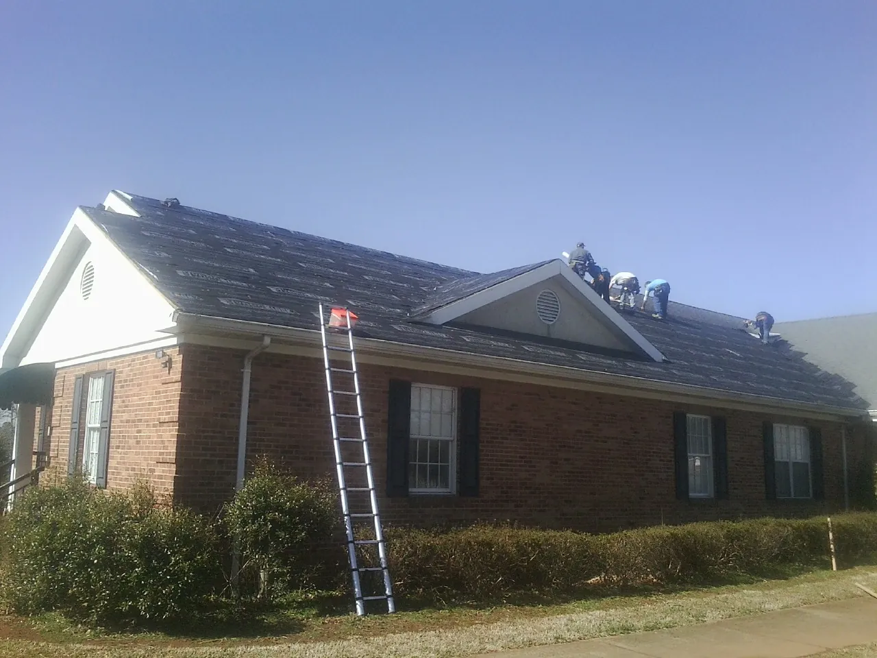 Crew tearing shingles off a brick ranch roof with a ladder set — Emergency Roof Repair work by our crew in Paul Smiths