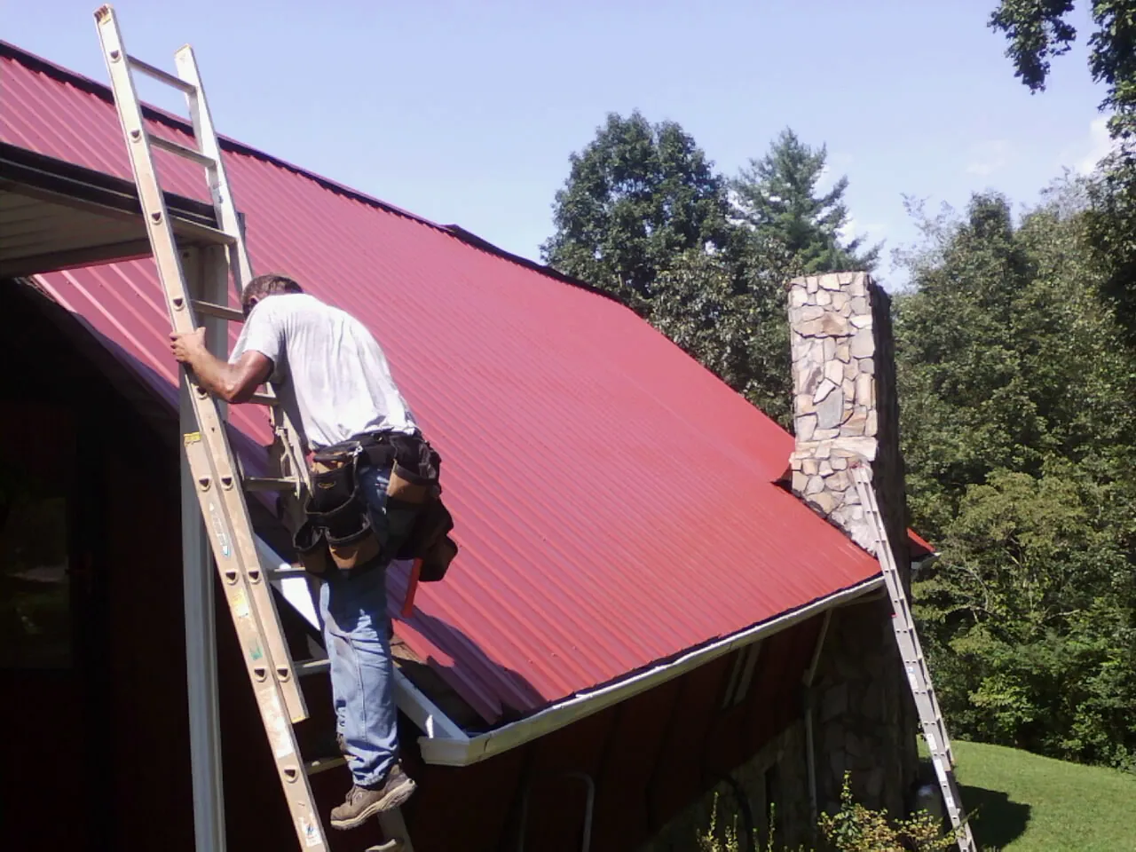 Roofer on a ladder beside a red metal roof and stone chimney — Soffit & Fascia Repair by Roofing Brothers in Paul Smiths
