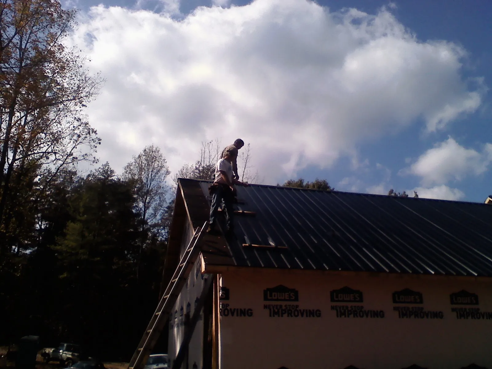 Roofer fastening dark metal panels high against a cloudy sky, from a Roof Inspection job in Paul Smiths